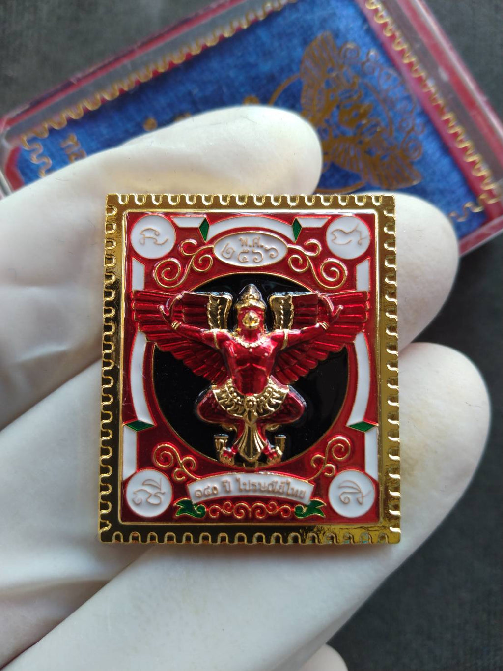 Garuda stamp medallion Thai Post ornate red and gold Thai amulet pendant with enamel details held in gloved hand