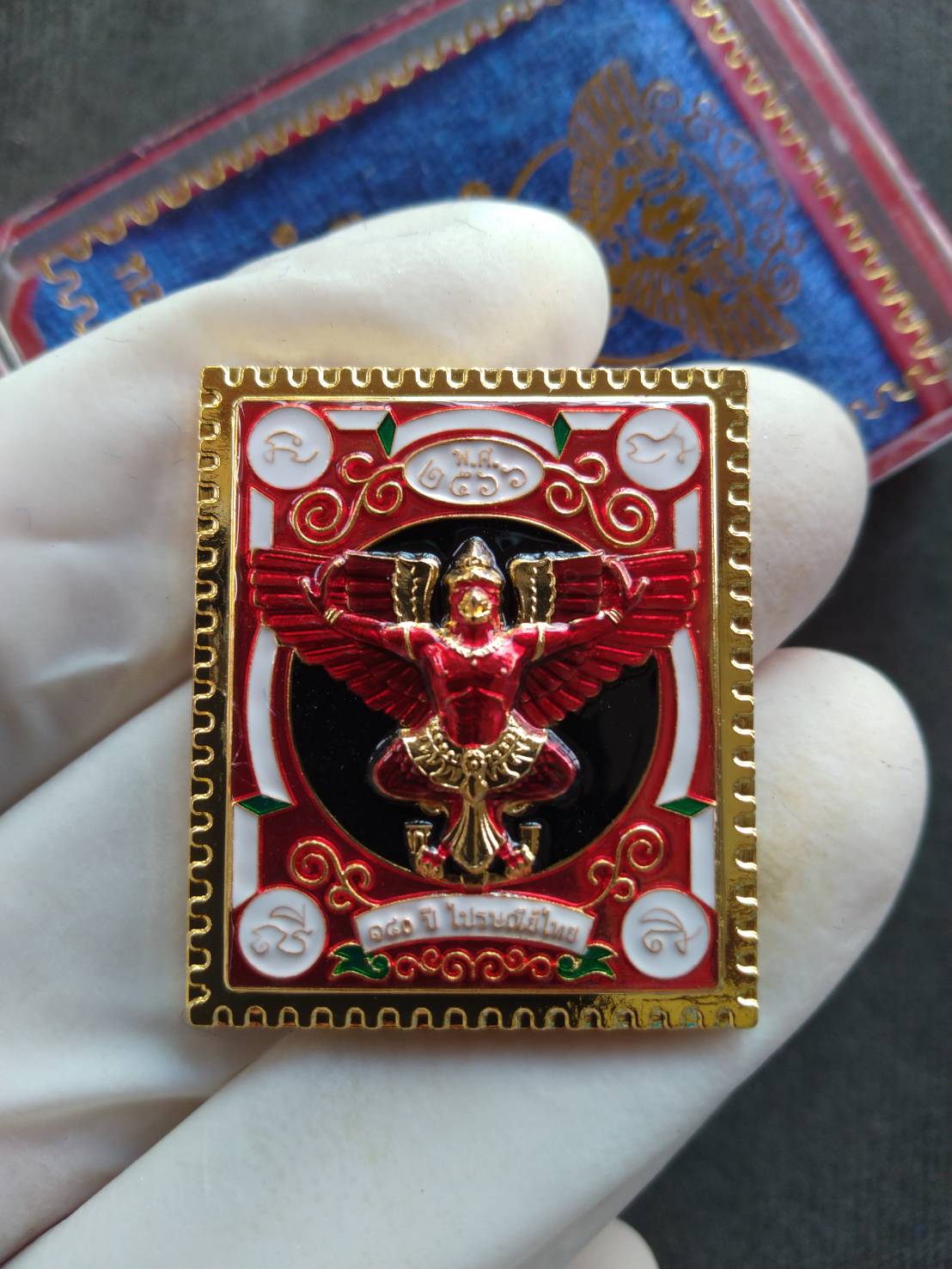 Garuda stamp medallion Thai Post ornate red and gold Thai amulet pendant with enamel details held in gloved hand
