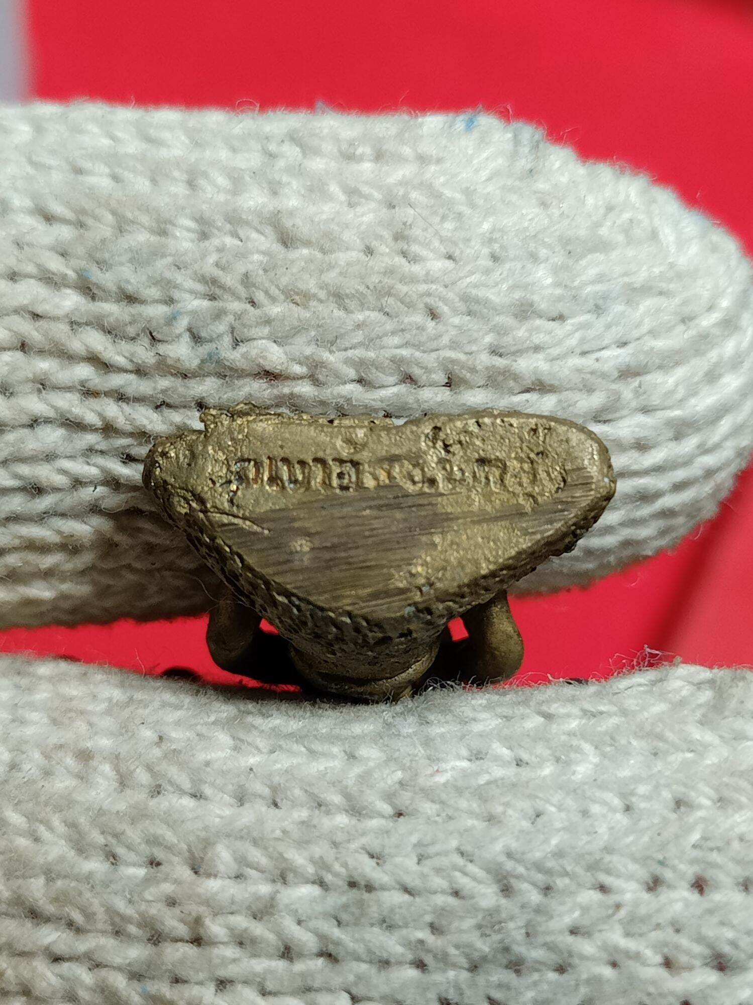 Close up of small bronze Ruesi Hermit Amulet Wat Khao Or showing engraved base held in white glove against red background