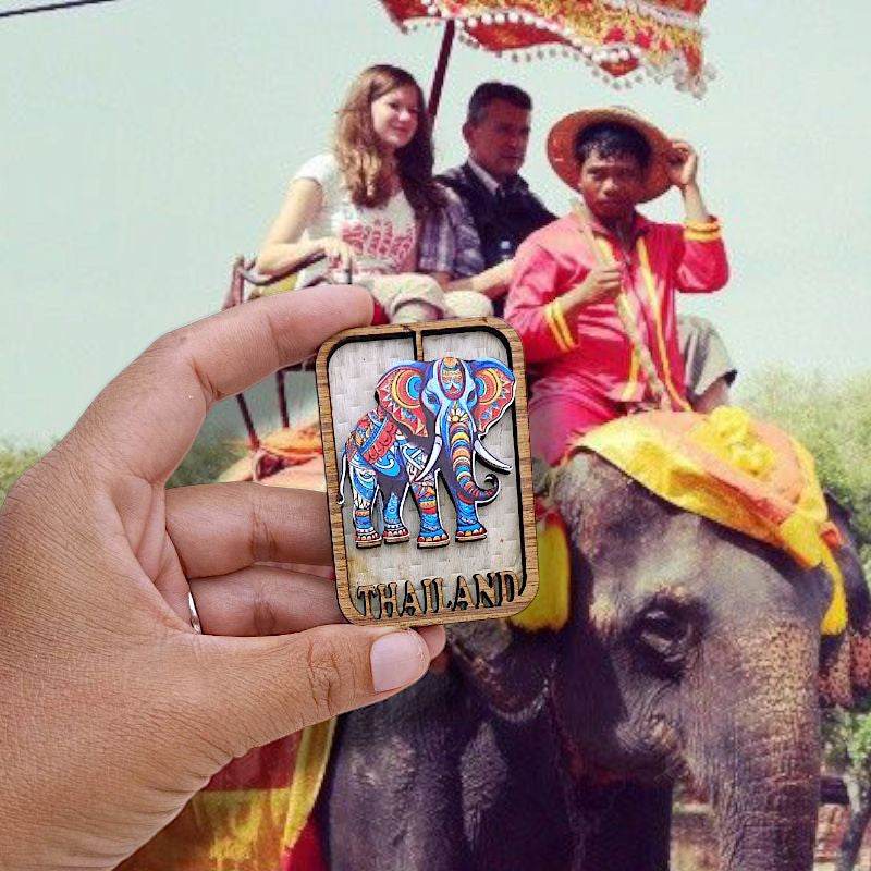 Hand holding colorful elephant Thailand souvenir magnets with tourists riding elephant in background