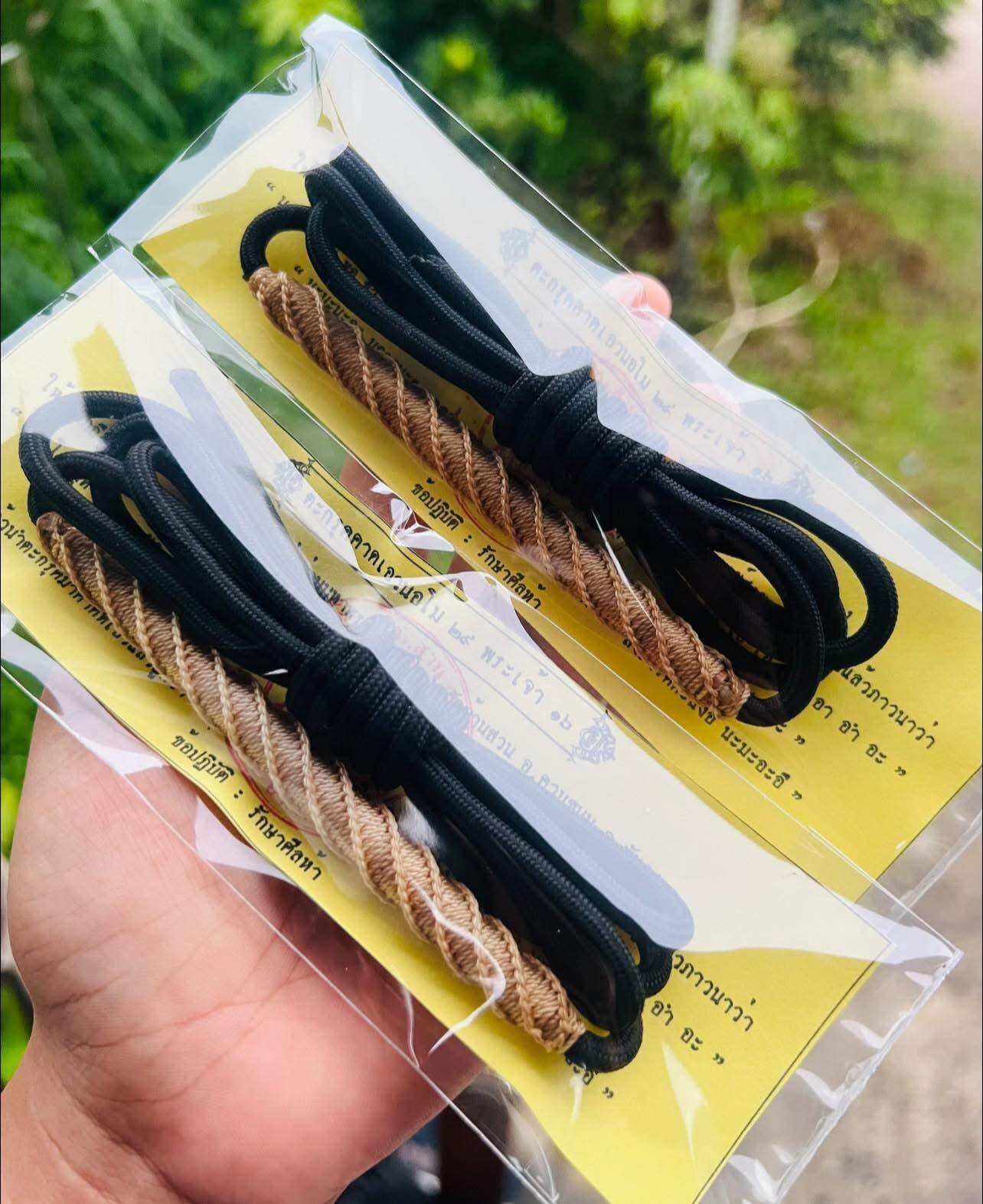 Two packaged braided gold and black waist cords on yellow card held in hand by Thai Blessing Takrud waist amulet for protection.