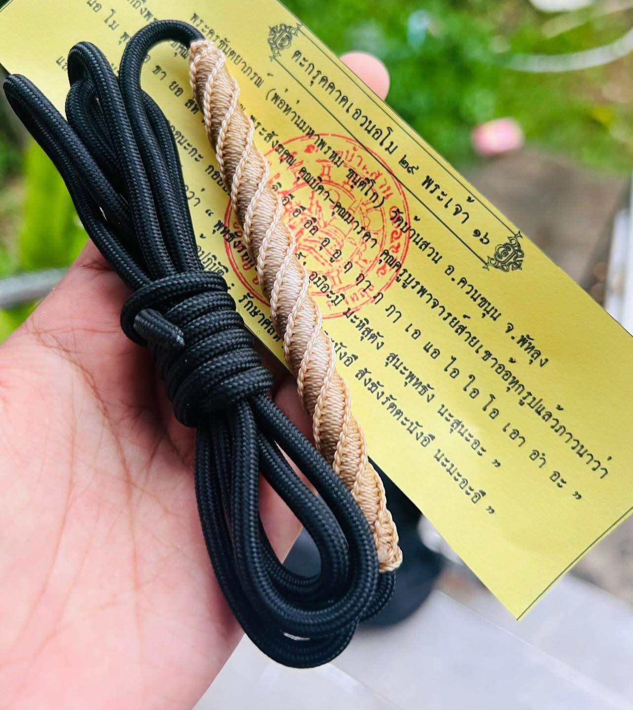 Hand holding Takrud waist amulet for protection braided gold cord with black waist rope and yellow Thai blessing card