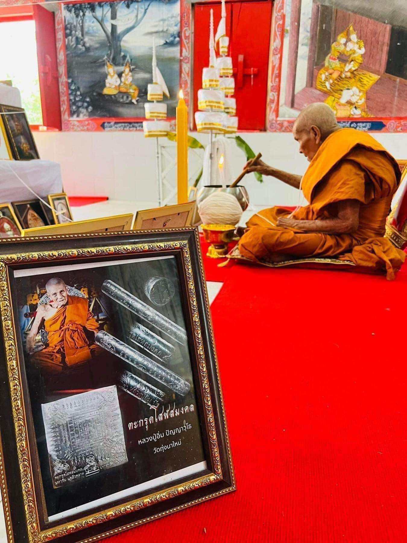 Takrut Solos Mongkol amulet displayed in framed poster while a monk performs a blessing ceremony
