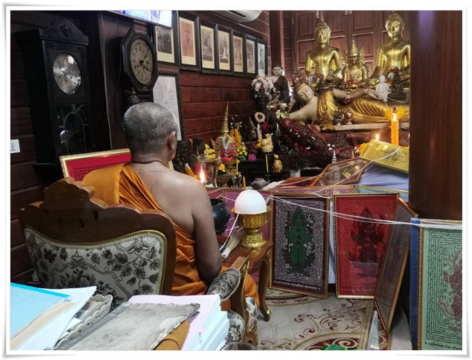 Monk seated in Thai temple beside altar with Buddha statues and hanging Thao Wessuwan yantra cloth panels