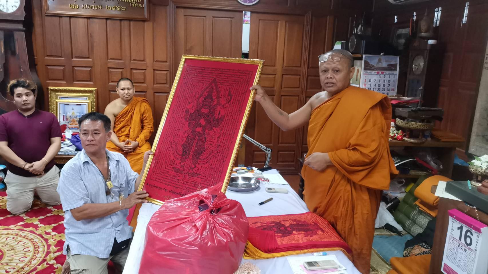 Monks and devotees in a Thai temple presenting a framed Thao Wessuwan yantra cloth red protective talisman
