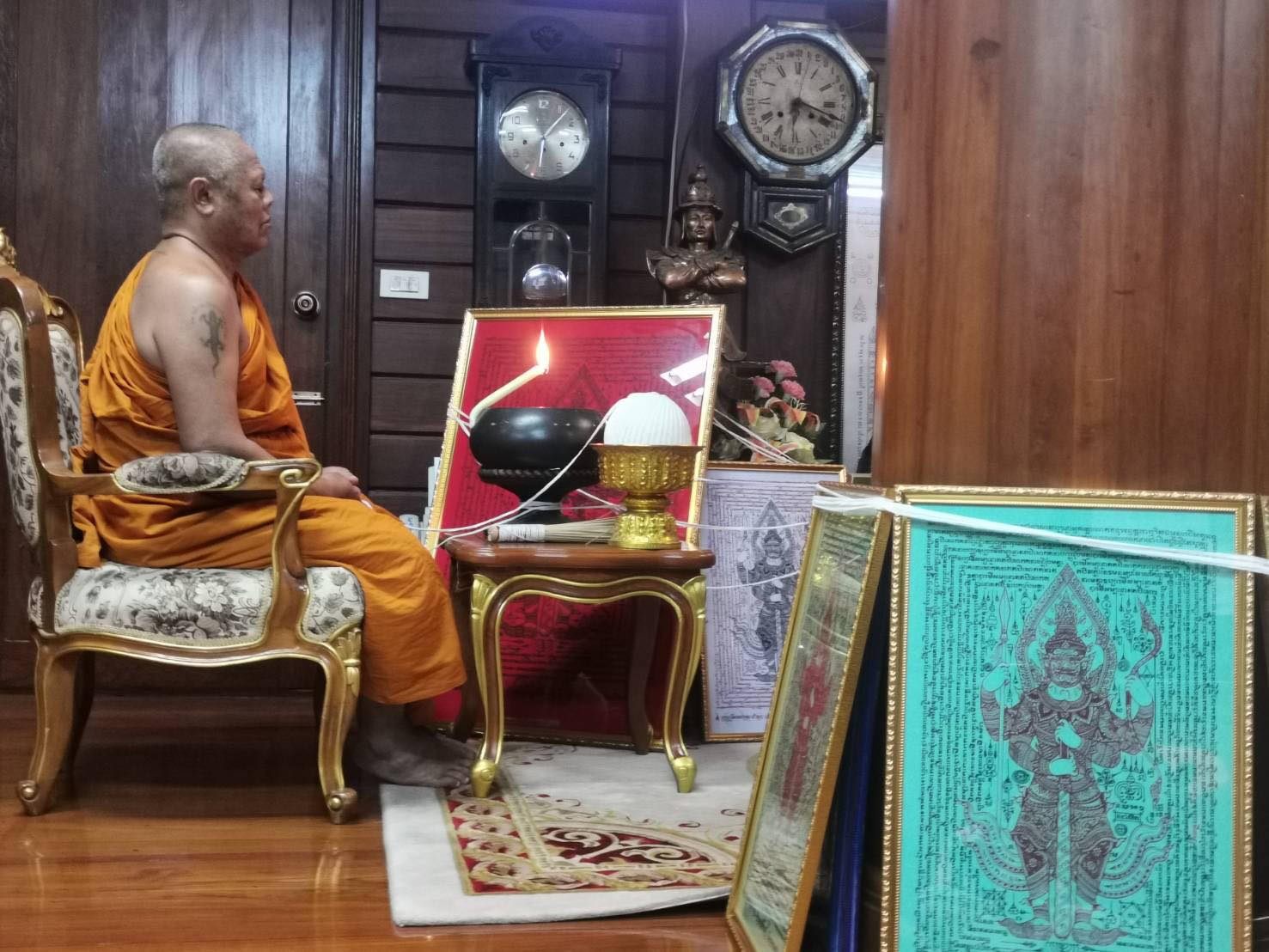 Buddhist monk with framed Thao Wessuwan yantra cloth and gold offering bowl in wooden shrine room