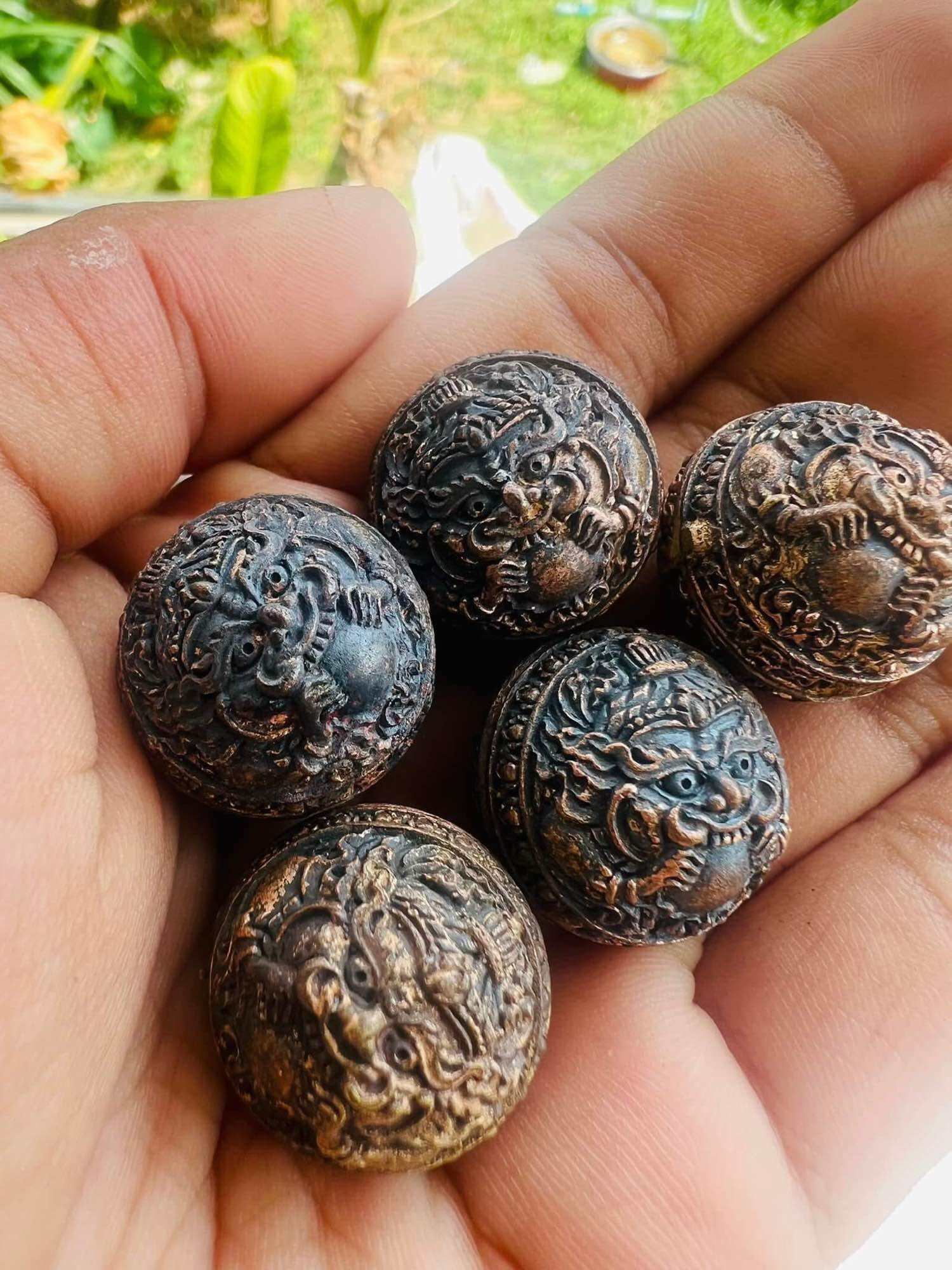 Hand holding five round bronze amulet beads with guardian face carvings ancient Thai talisman first edition