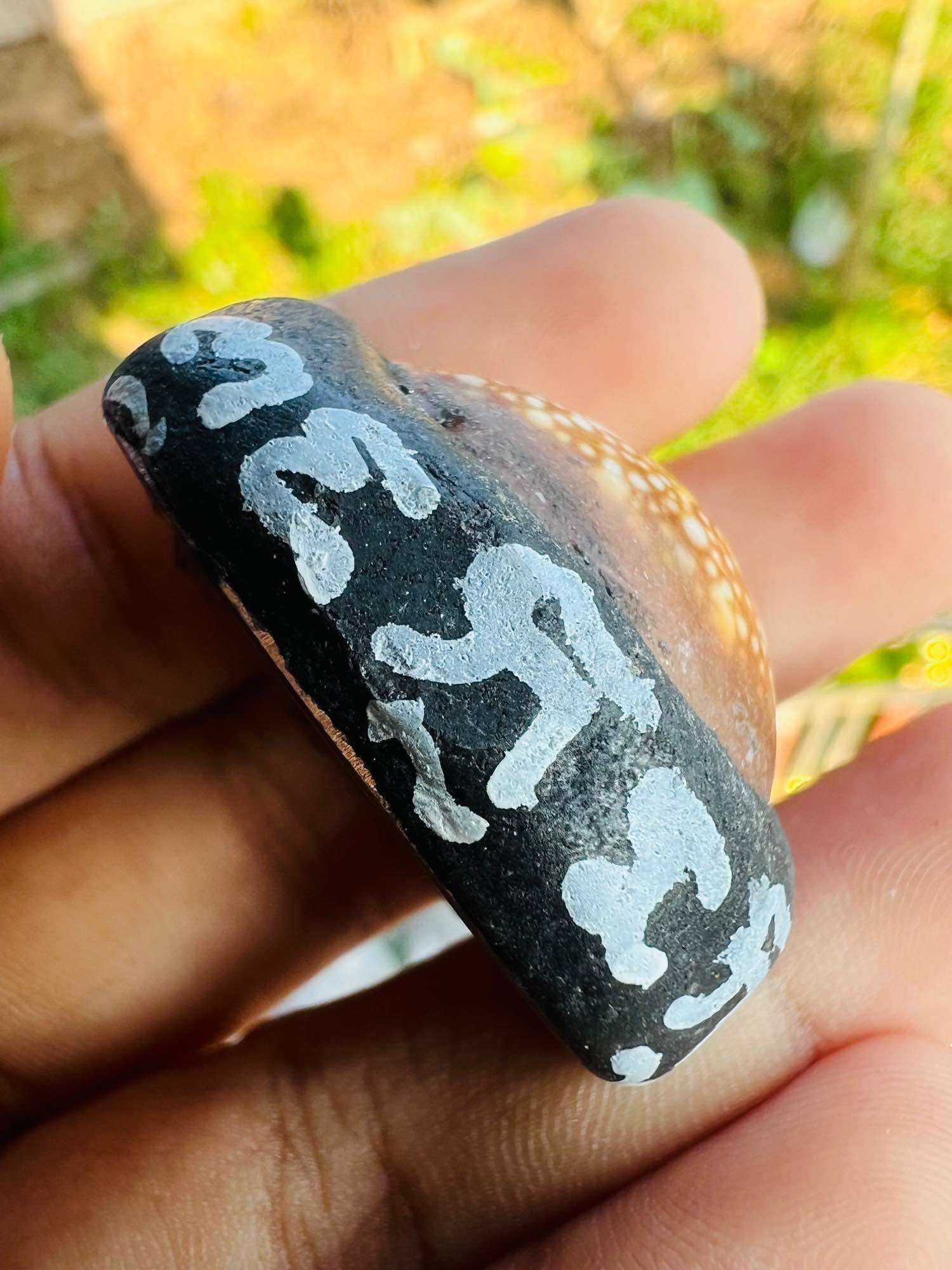 Hand holding Thai shell amulet with painted white script on black edge, Bia Gae amulet for protection.