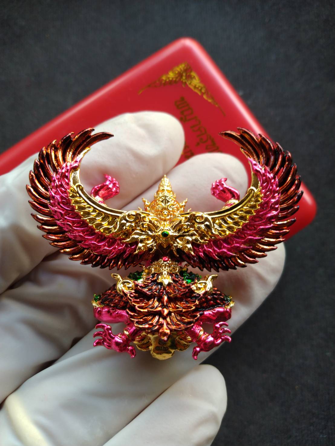 Gloved hand holding gold and pink Garuda Phaya Krut Yok Tana Amulet Garuda Wealthy Millionaire Edition ornate winged Thai amulet