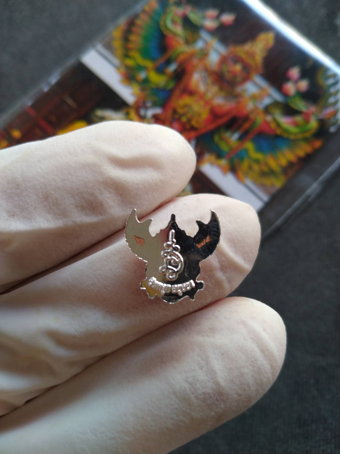 Thai Blessing Co mini Garuda amulet Thailand metal pendant held by gloved fingers with colorful Garuda card background