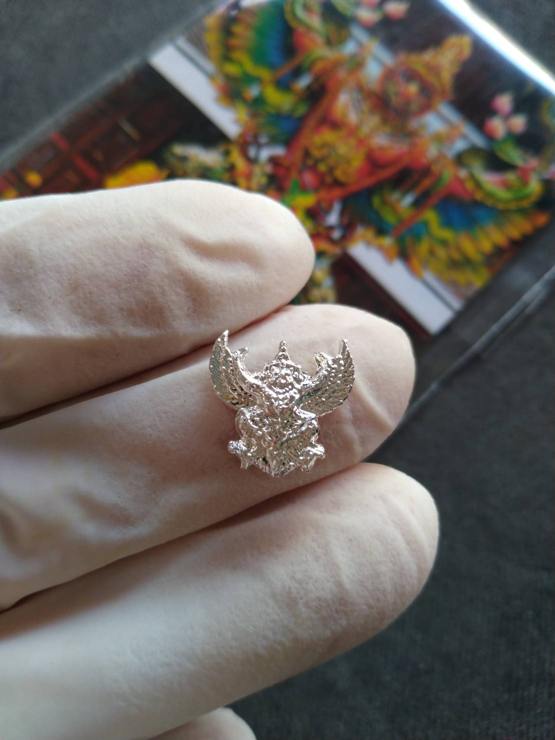 Silver mini Garuda amulet Thailand pendant held by gloved fingers with colorful Garuda card