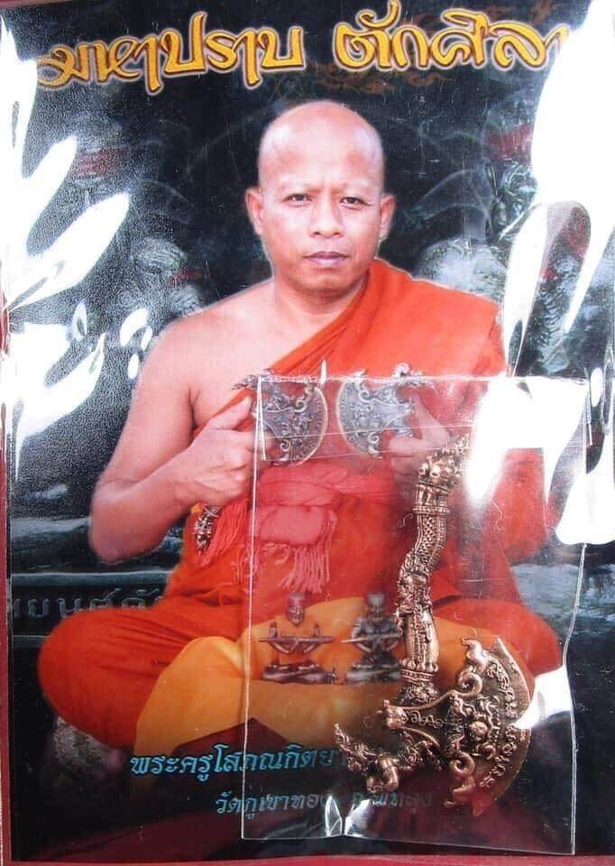 Phaya Khwan Maha Prap Amulet ornate metal amulet displayed over photo of seated Buddhist monk in orange robes