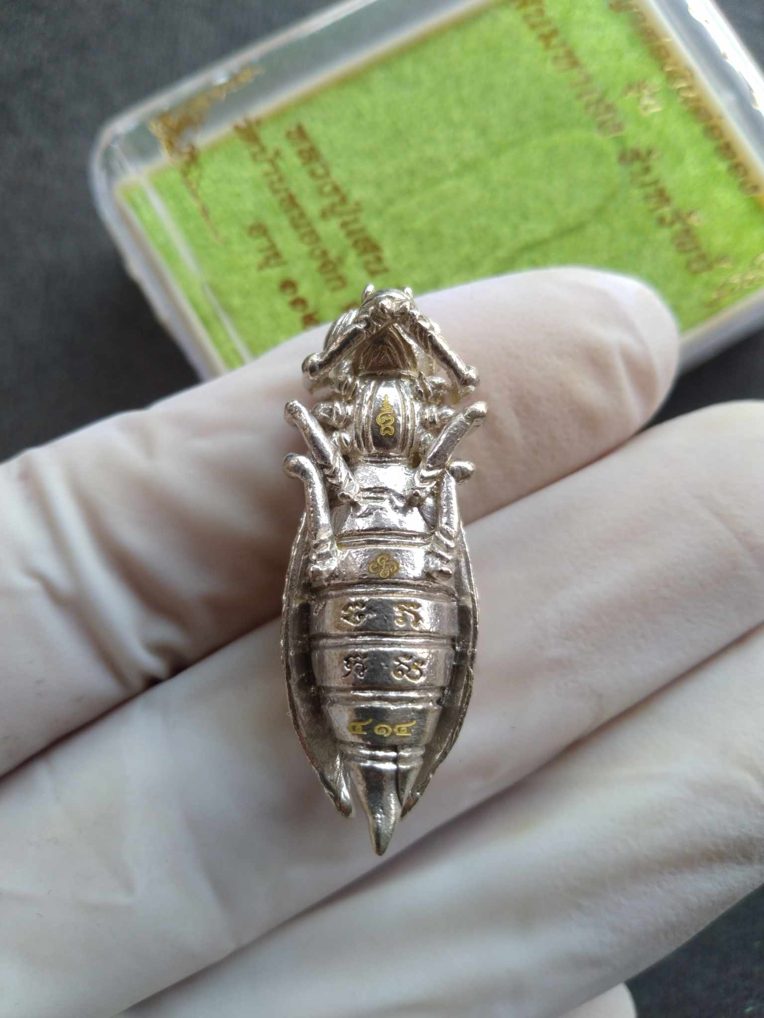 Phaya Tor Ngern Tor Thong amulet silver insect style pendant with gold script held in gloved hand