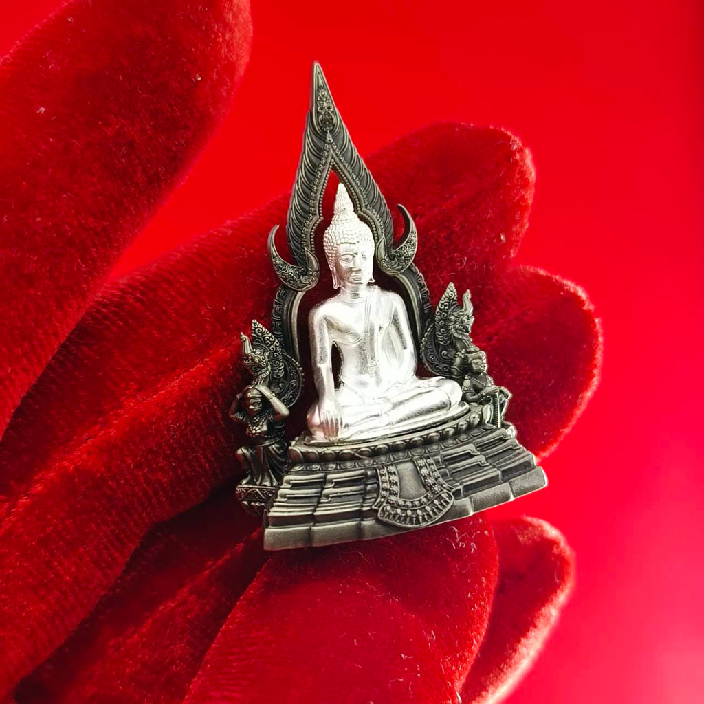 Silver Phra Phuttha Chinnarat amulet seated Buddha on ornamental base held on red velvet background
