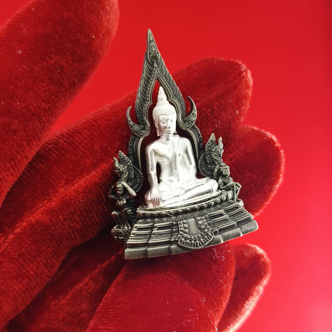 Silver Phra Phuttha Chinnarat amulet seated Buddha on ornamental base held on red velvet background