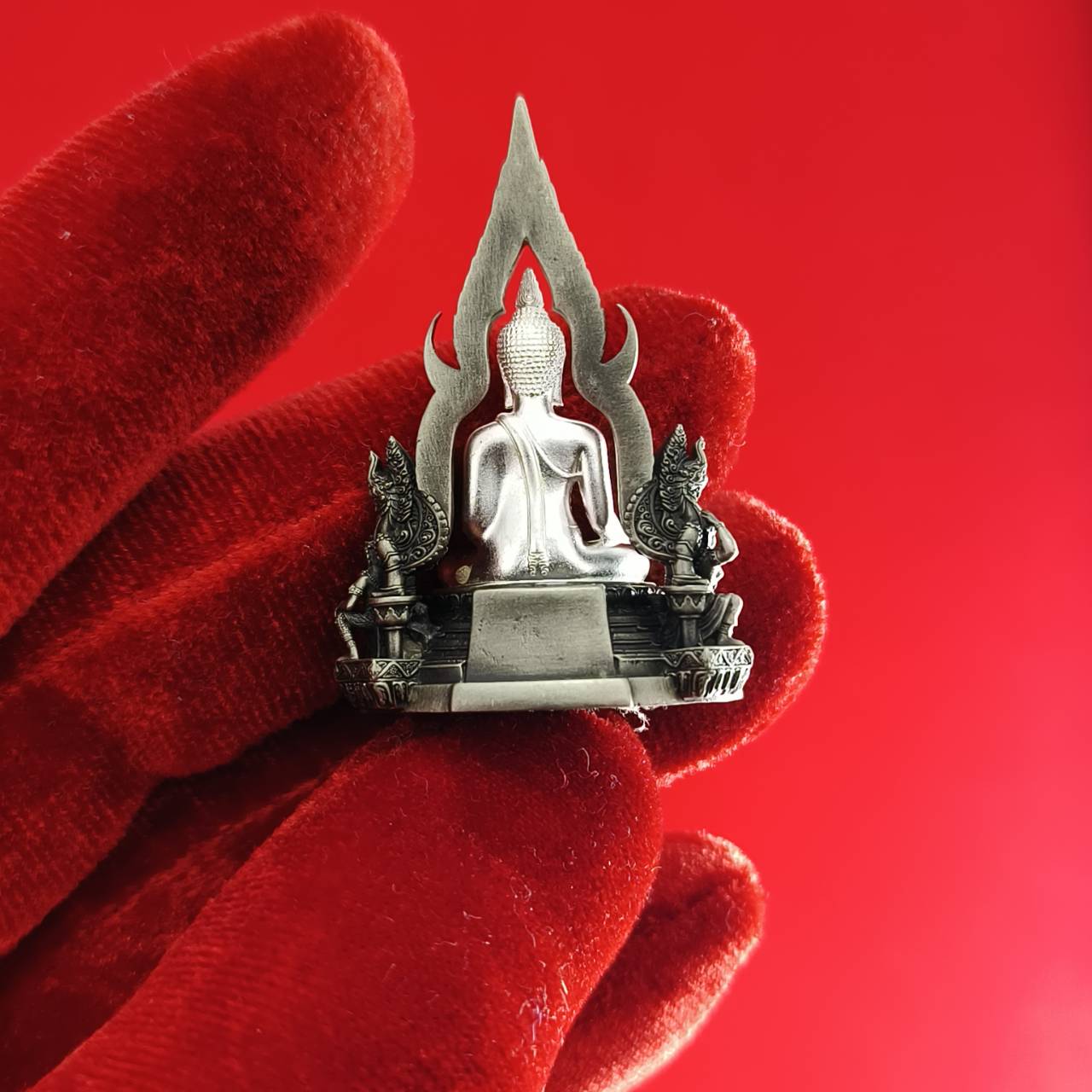 Silver Phra Phuttha Chinnarat amulet held on red velvet glove detailed Thai Buddha shrine design