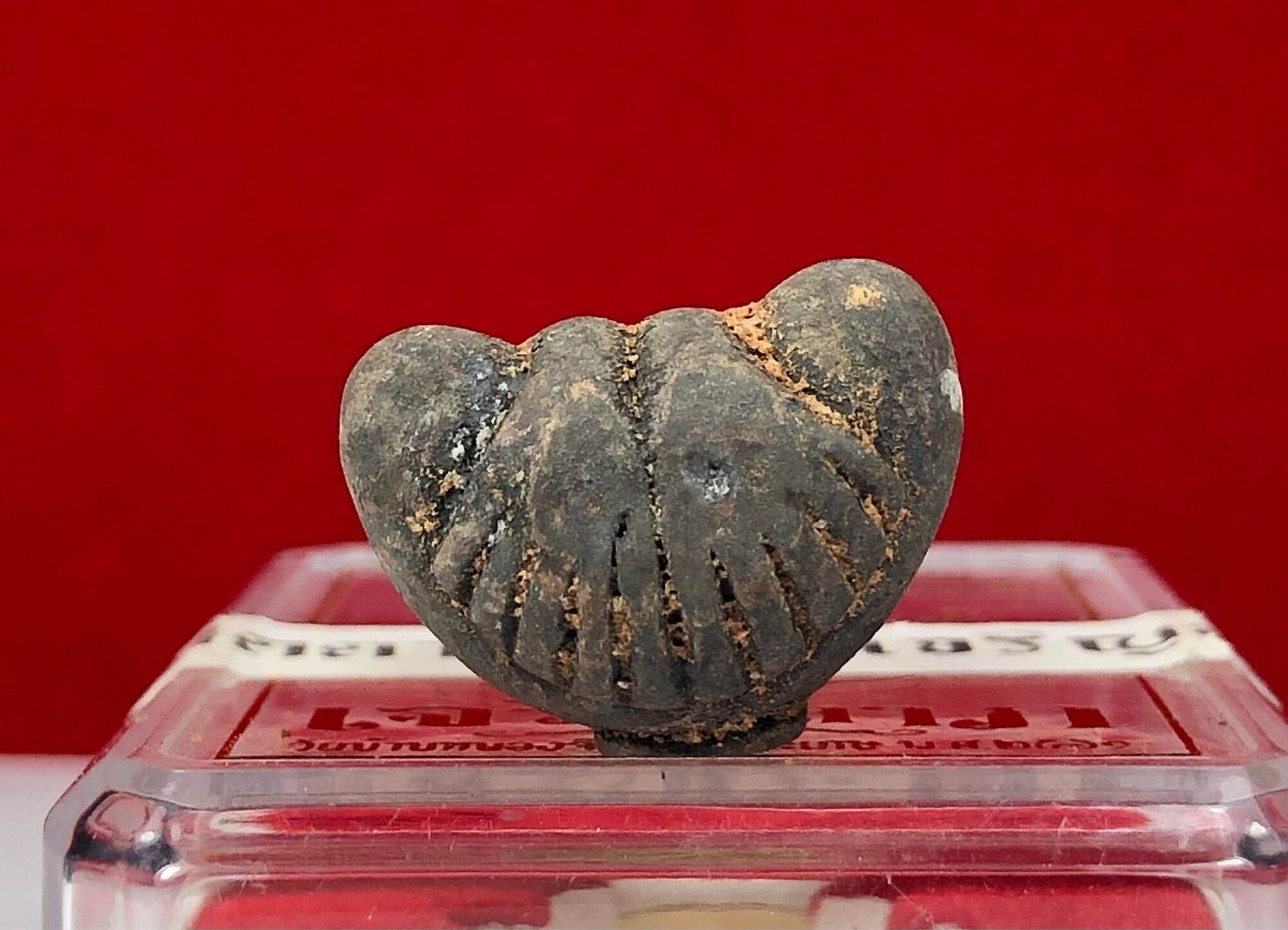 Phra Pidta Ancient Amulet small worn dark clay pendant on clear display box against red background