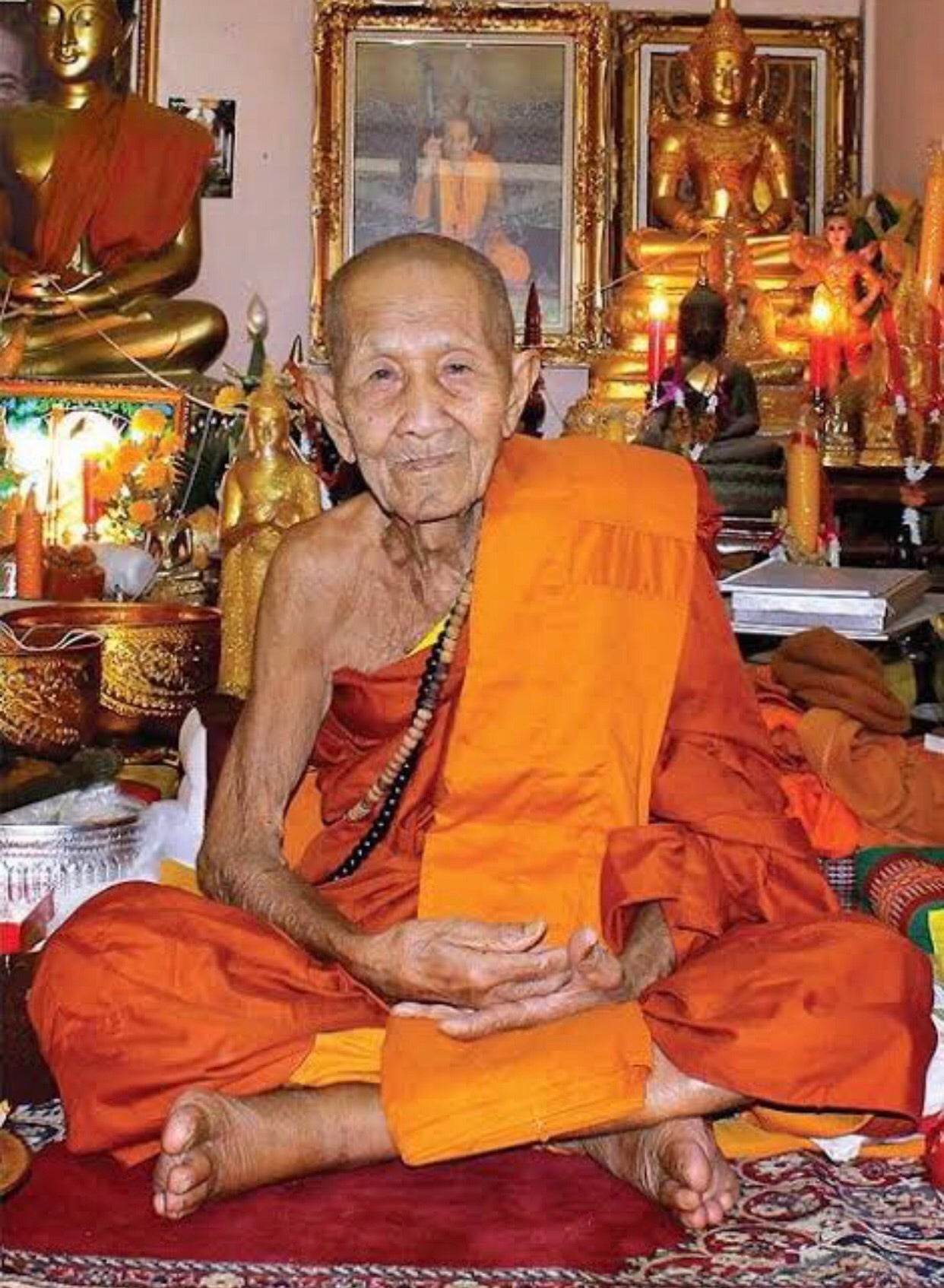 Phra Pidta Ancient Amulet shown with elderly Buddhist monk seated in shrine among Buddha statues