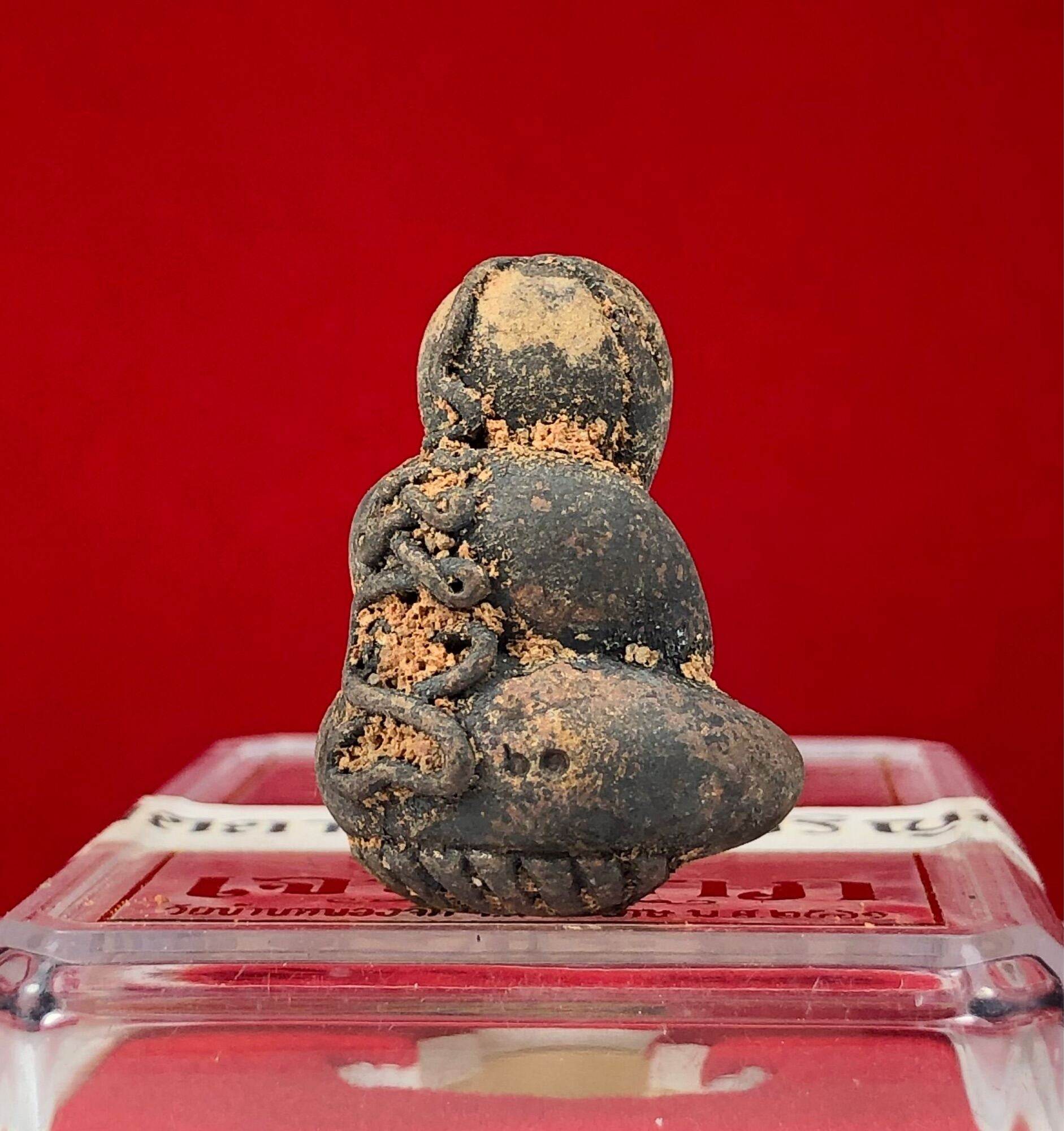 Phra Pidta Ancient Amulet dark clay amulet with dirt encrustation on clear display case red background