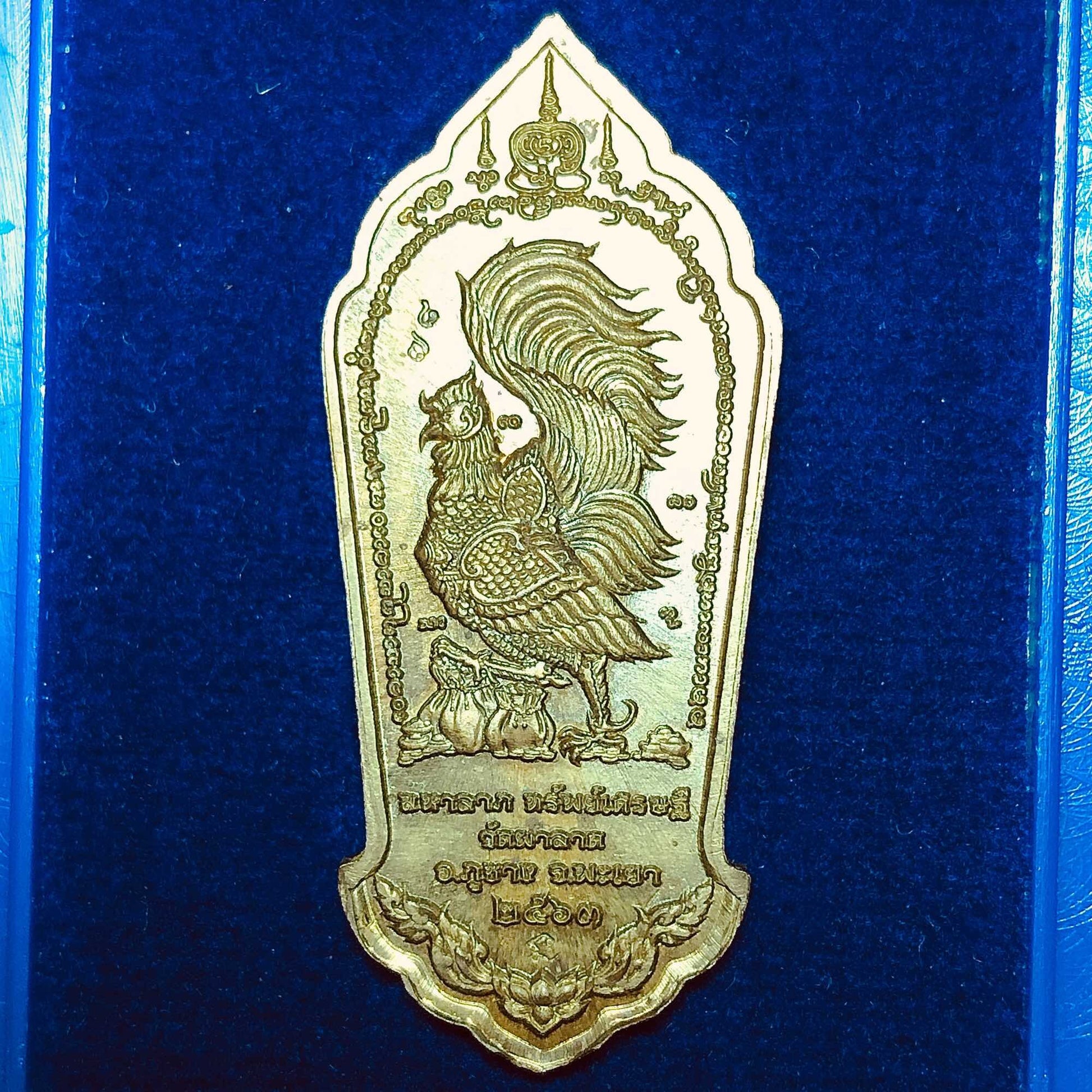 Gold Thai metal pendant engraved rooster on blue velvet display Phra Pidta Maha Lap amulet for wealth.