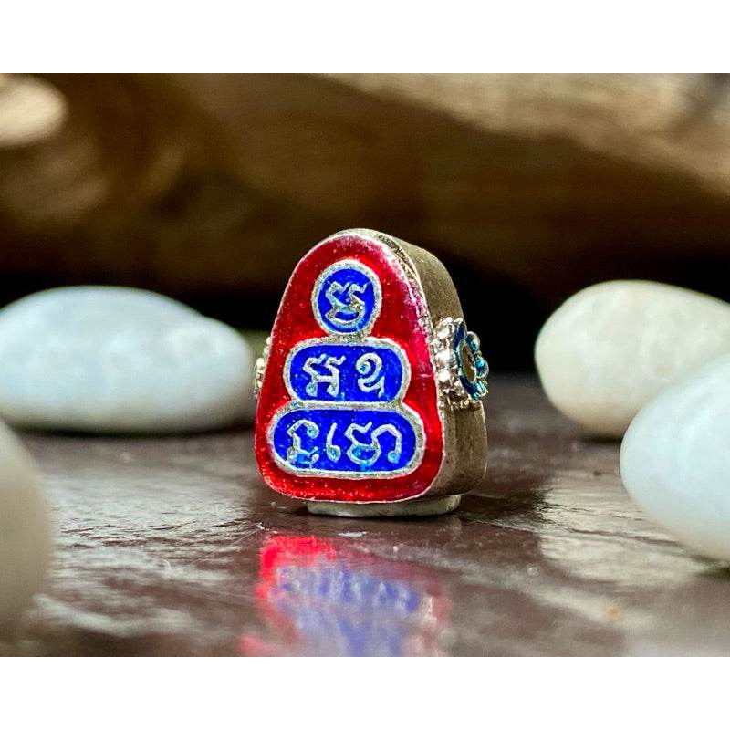 Phra Pidta wealth amulet red and blue enamel Thai script metal charm on wooden surface with white pebbles