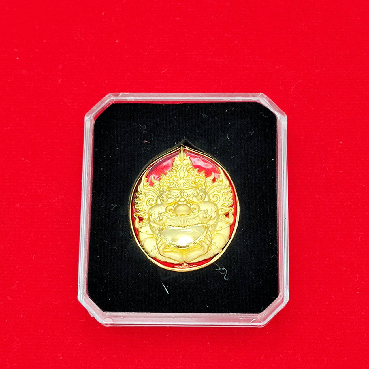 Ornate gold Rahu deity amulet medallion in clear display case on black velvet with red background