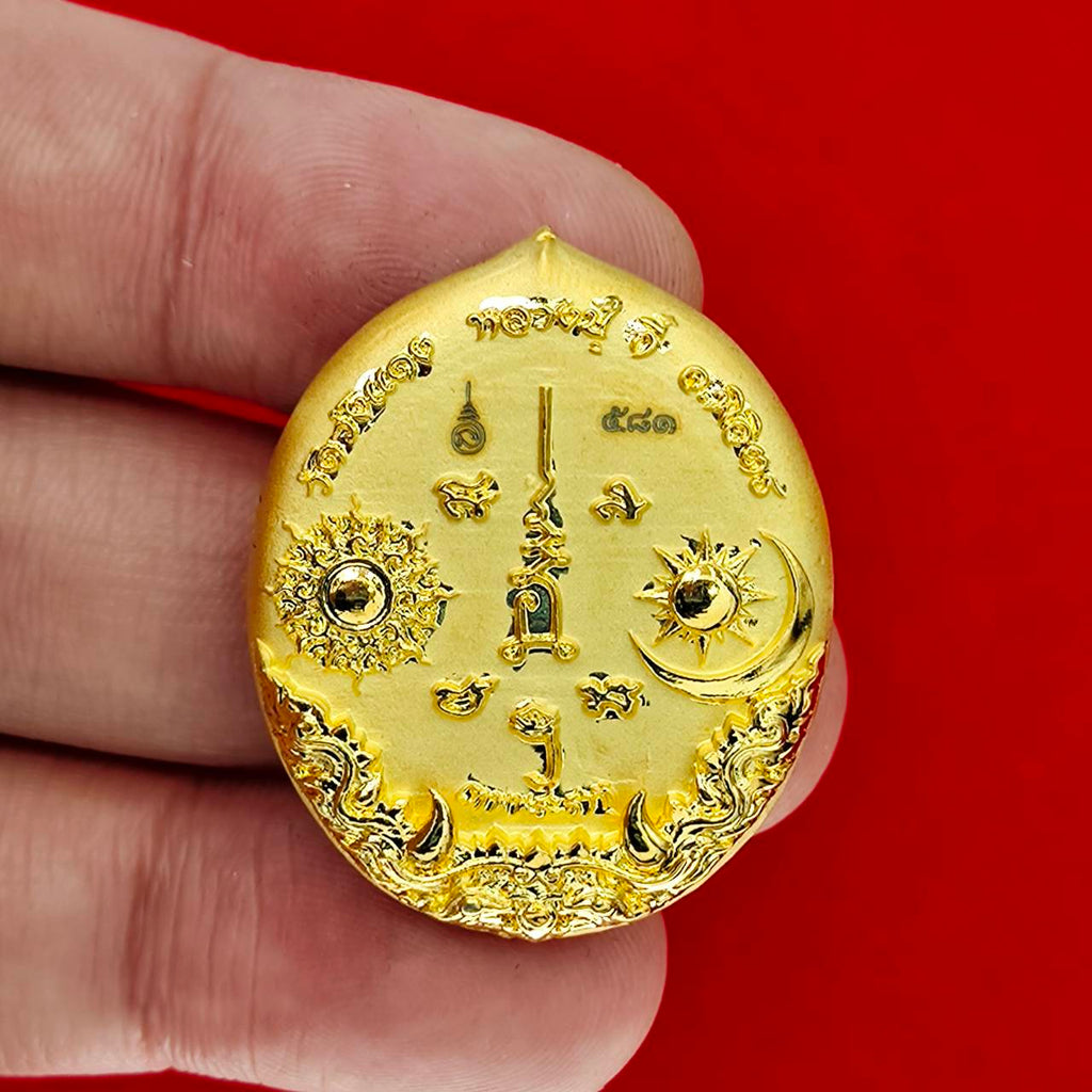 Gold oval Rahu deity amulet with sun and moon motifs and Thai script held between fingers on red background