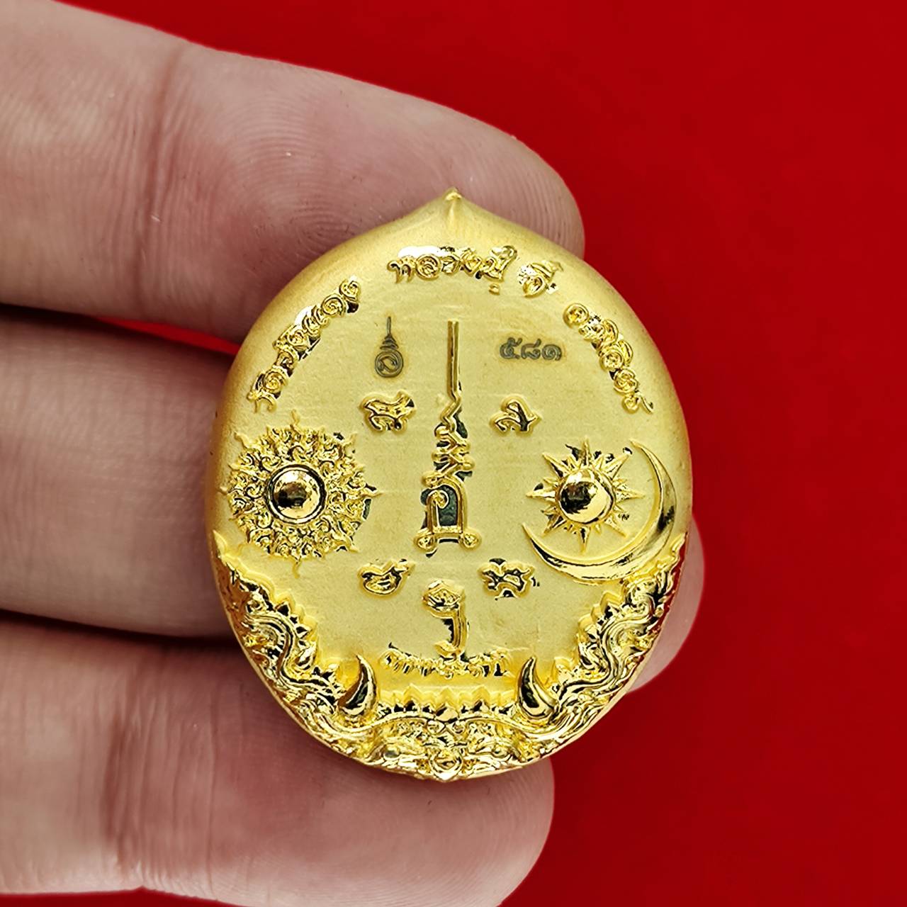 Gold oval Rahu deity amulet with sun and moon motifs and Thai script held between fingers