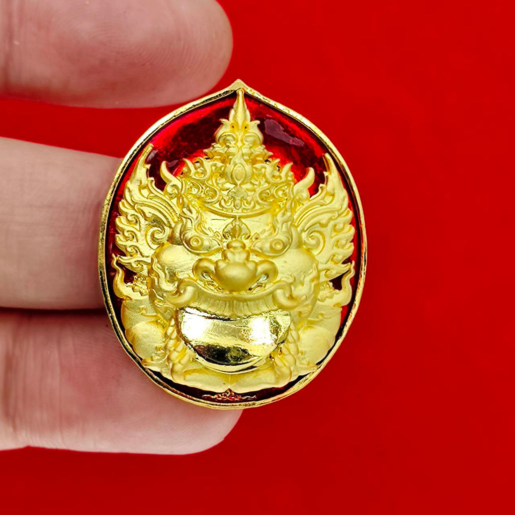 Rahu deity amulet gold enamel medallion with intricate face on red background held between fingers