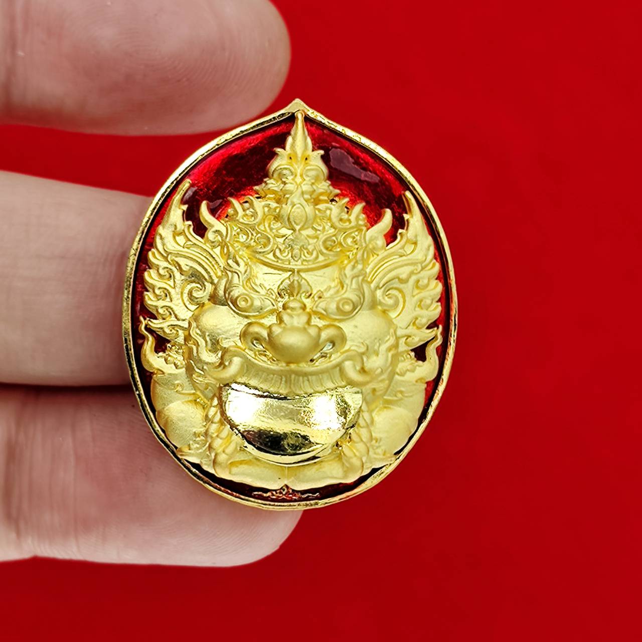 Rahu deity amulet gold enamel medallion with intricate face on red background held between fingers