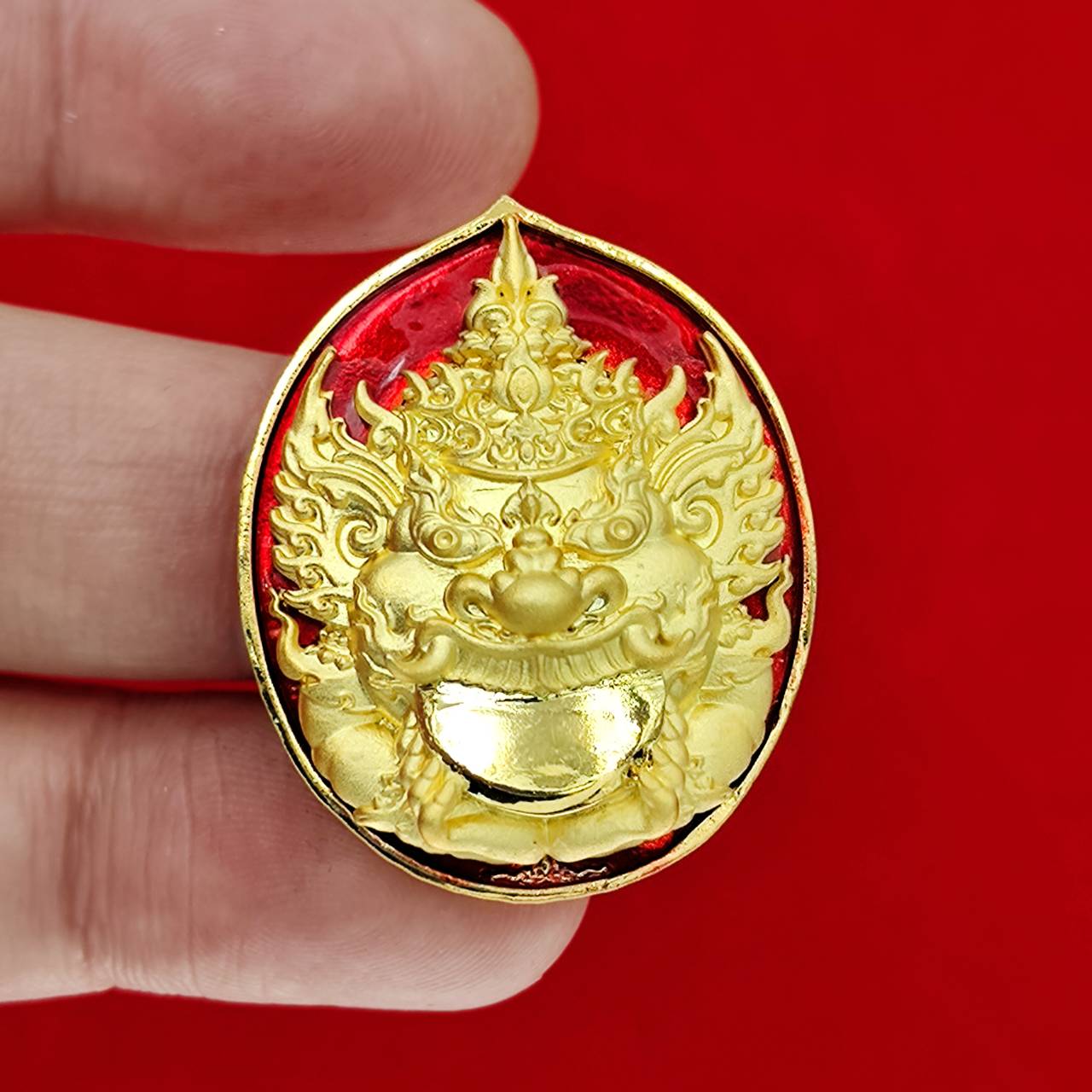 Rahu deity amulet gold medallion with ornate demon face and red enamel background held between fingers