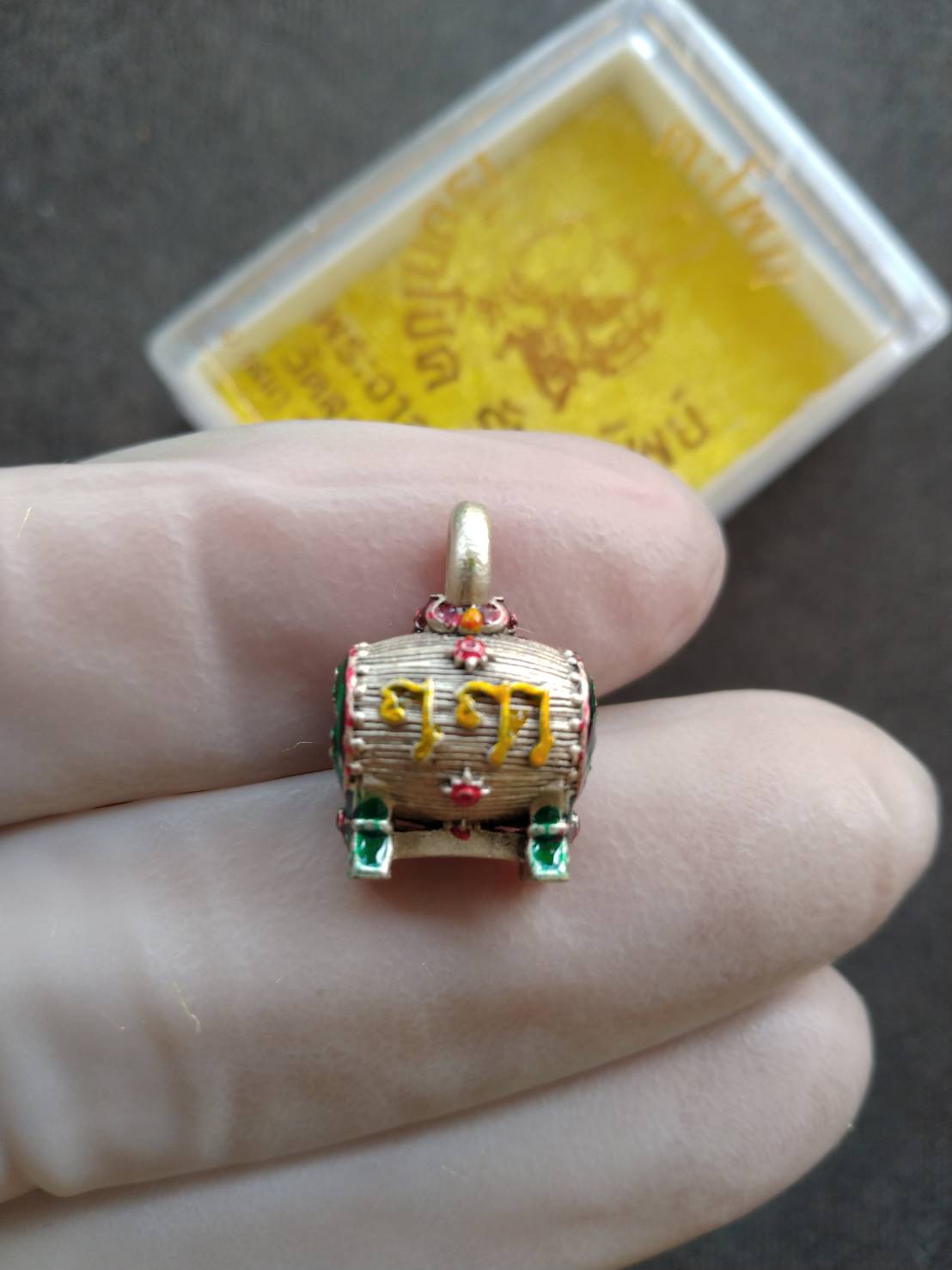 Sacred Drum Amulet for Wealth Prosperity Blessed by Phra Ajarn Tao. Gold drum pendant with red green enamel held in gloved hand