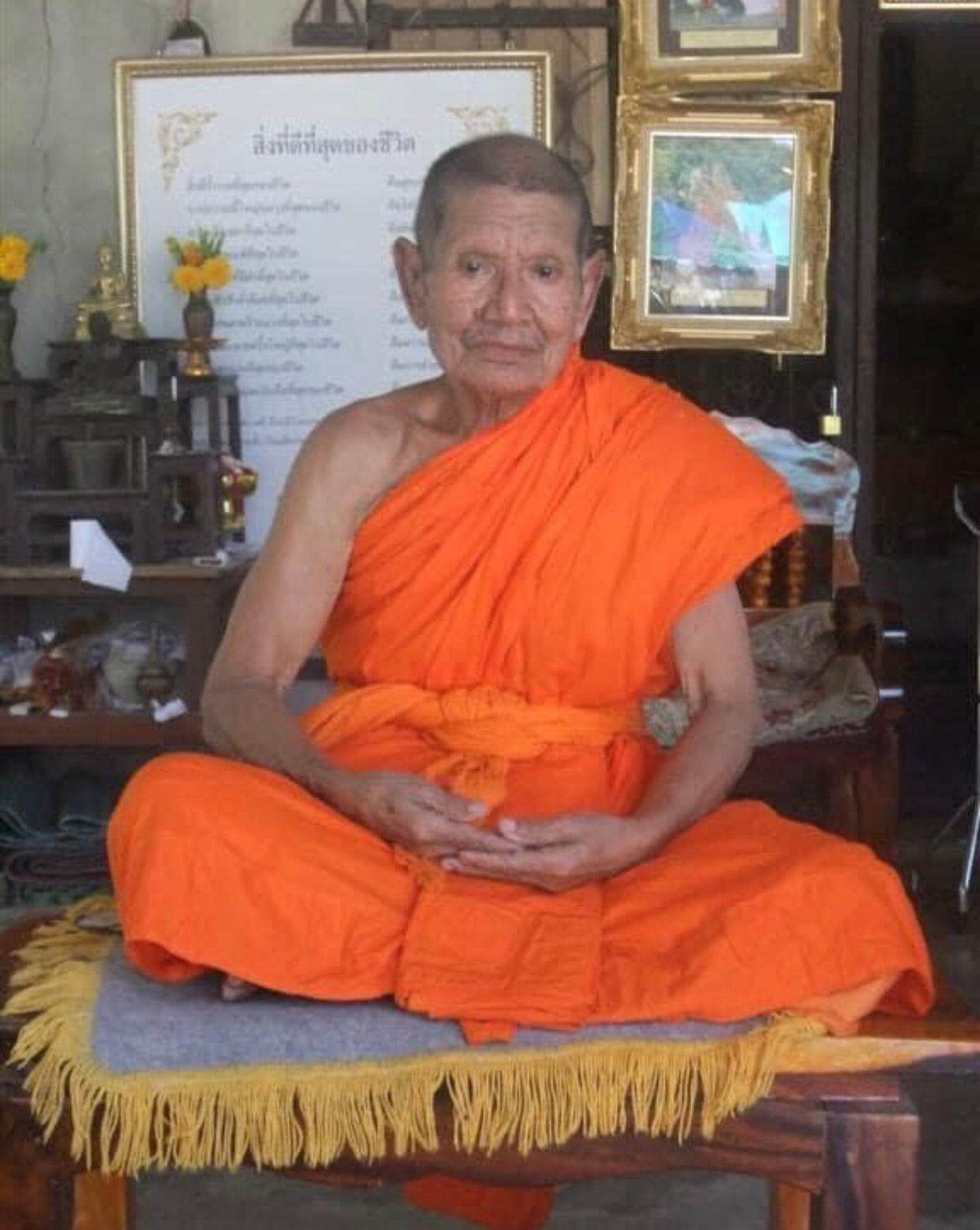 Elder Thai monk in orange robes seated at a home shrine offering Sarika couple amulet for love pendant blessing