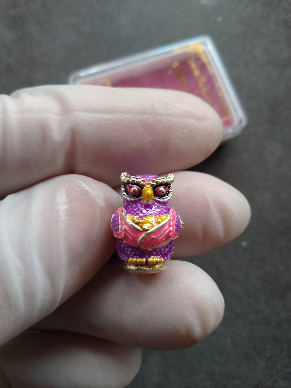 Close up of a small purple owl amulet held by gloved fingers Thep Rattikan oom sap amulet with gold and red detailing