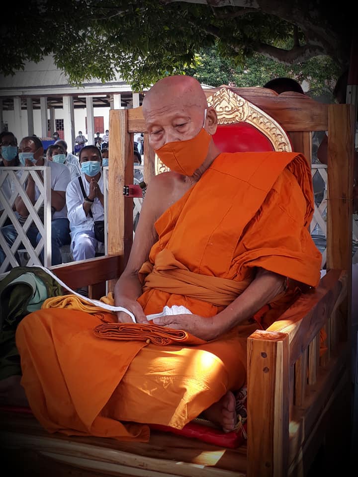 Monk in orange robes holding Thao Wessuwan yantra cloth during a serene ceremony outdoors