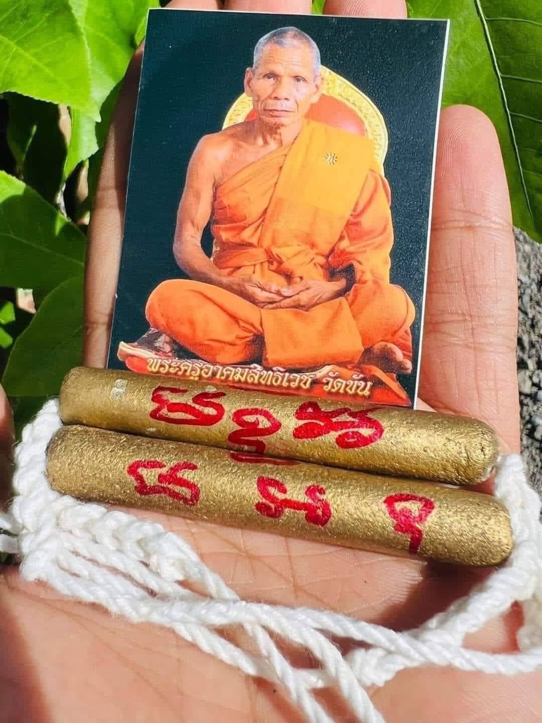 Hand holding laminated monk photo with two gold amulet sticks and white cord authentic Thai spiritual talisman