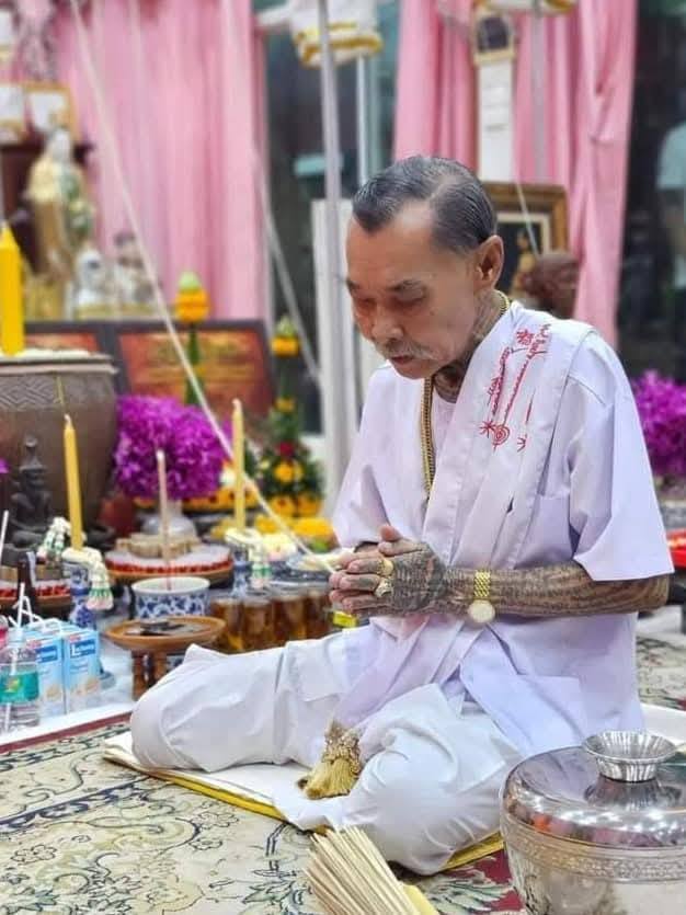 Tattooed elder performing Thai blessing ritual at home altar, blessing a one-eyed Rahu Thai amulet for protection and luck