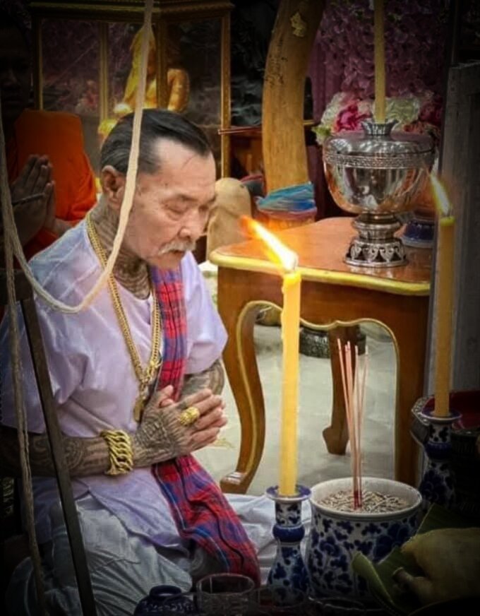 Elderly worshipper at shrine with candles and incense wearing a one-eyed Rahu Thai amulet pendant by Thai Blessing Co