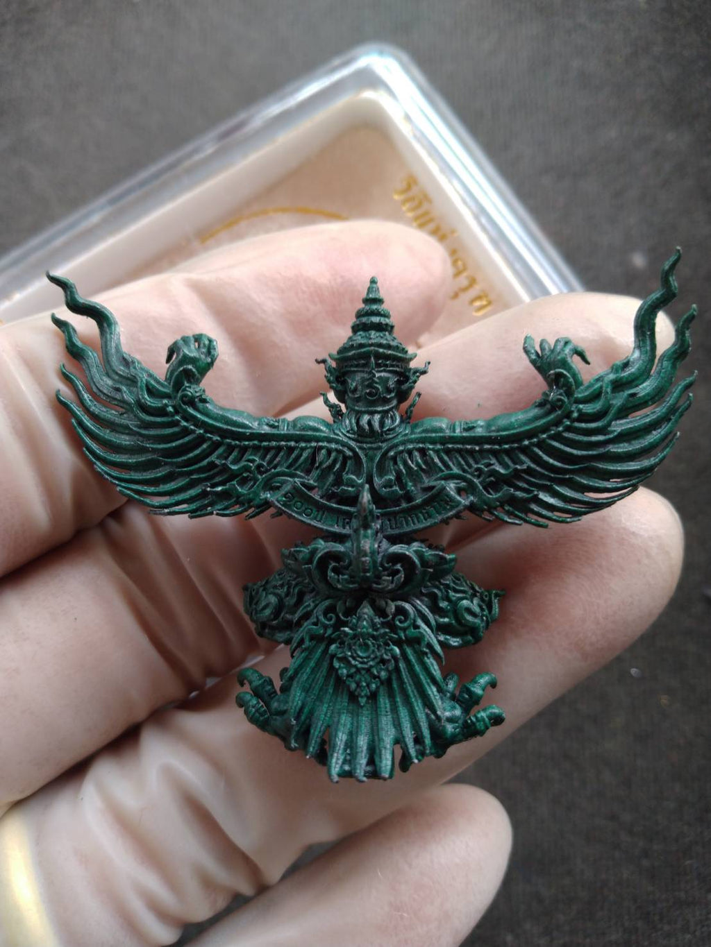 Hand holding green metal Phaya Krut amulet blessed by Luang Phor Phat. Intricate Garuda pendant from Thai Blessing Co