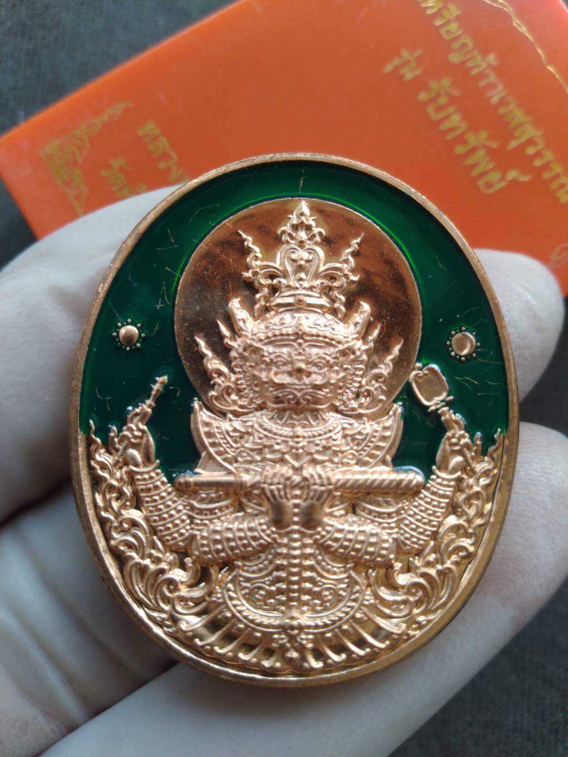 Thao Wessuwan amulet for protection gold oval pendant with green enamel background held by gloved hand