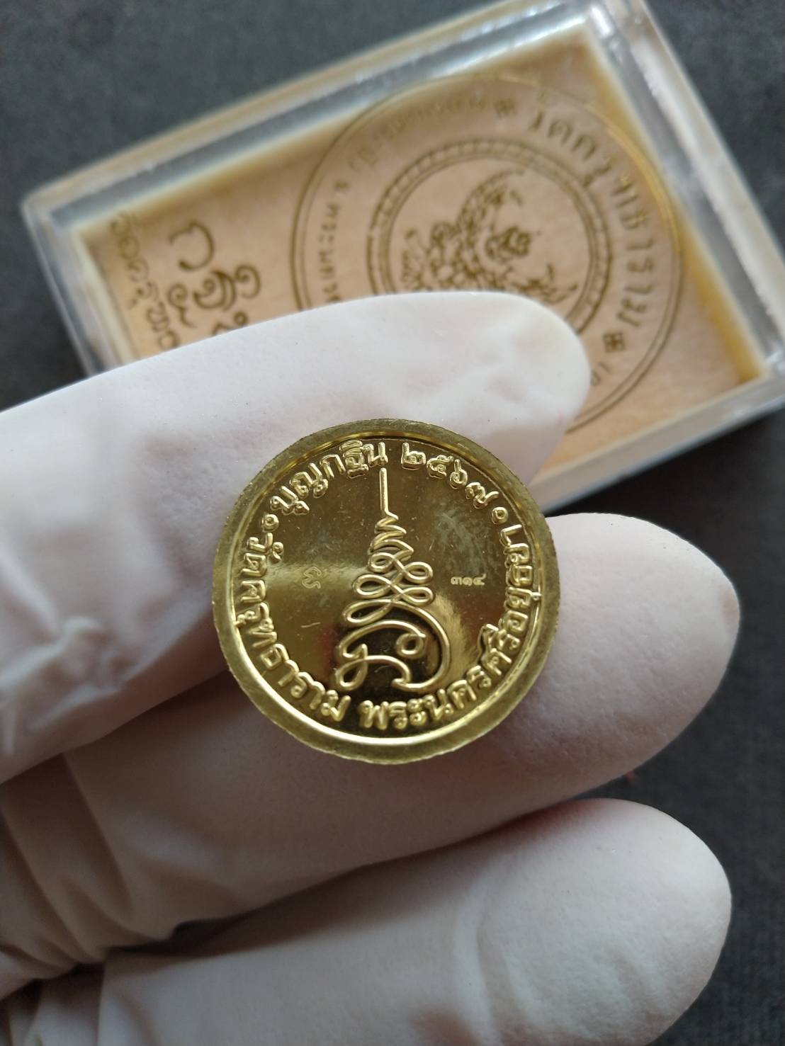 Gloved hand holding gold Garuda protection medallion with Thai script and clear display box by Thai Blessing Co