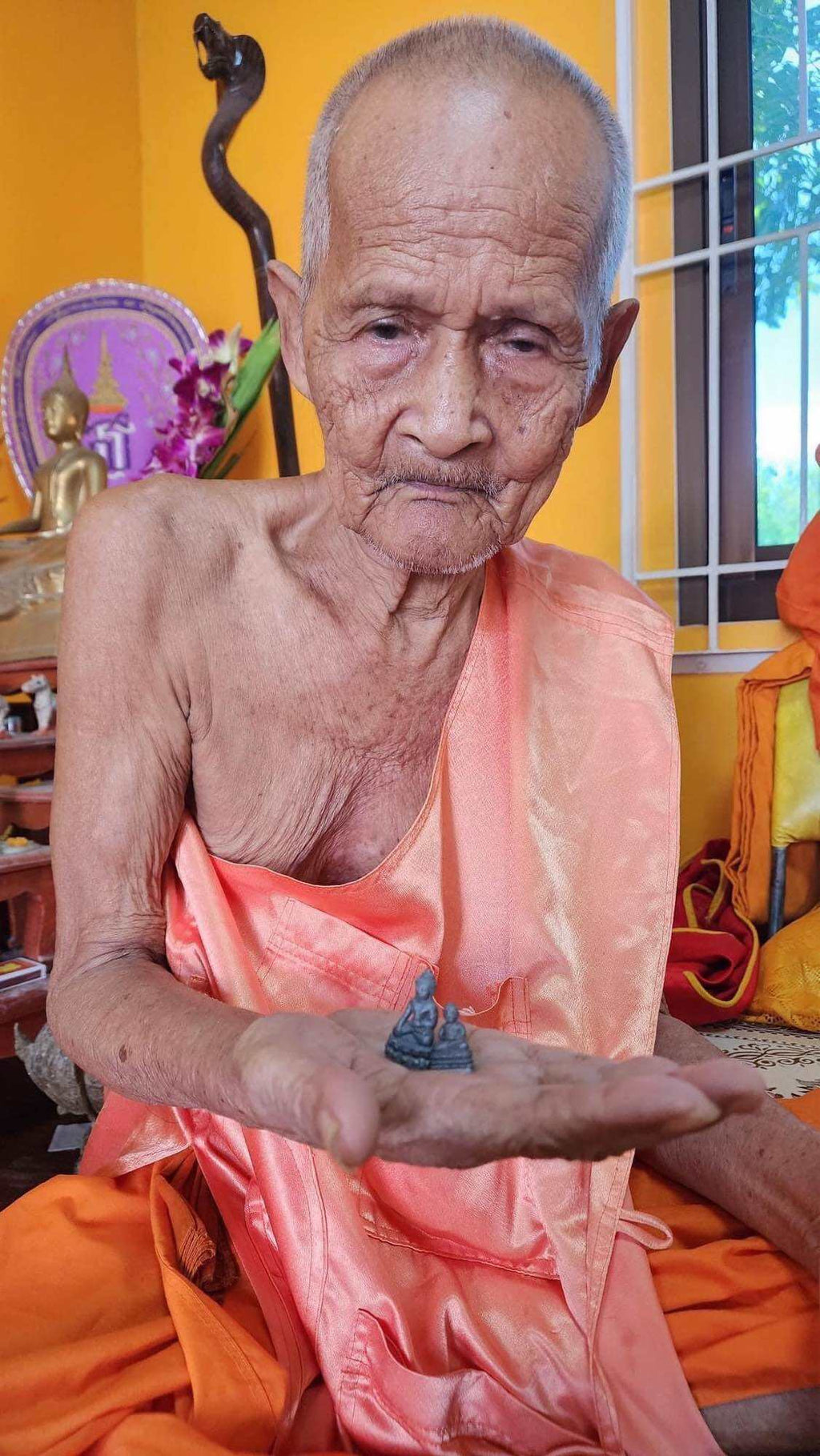 Elder Thai monk in orange robe holding Mongkol Hemarat Kring spiritual charm amulet in temple setting