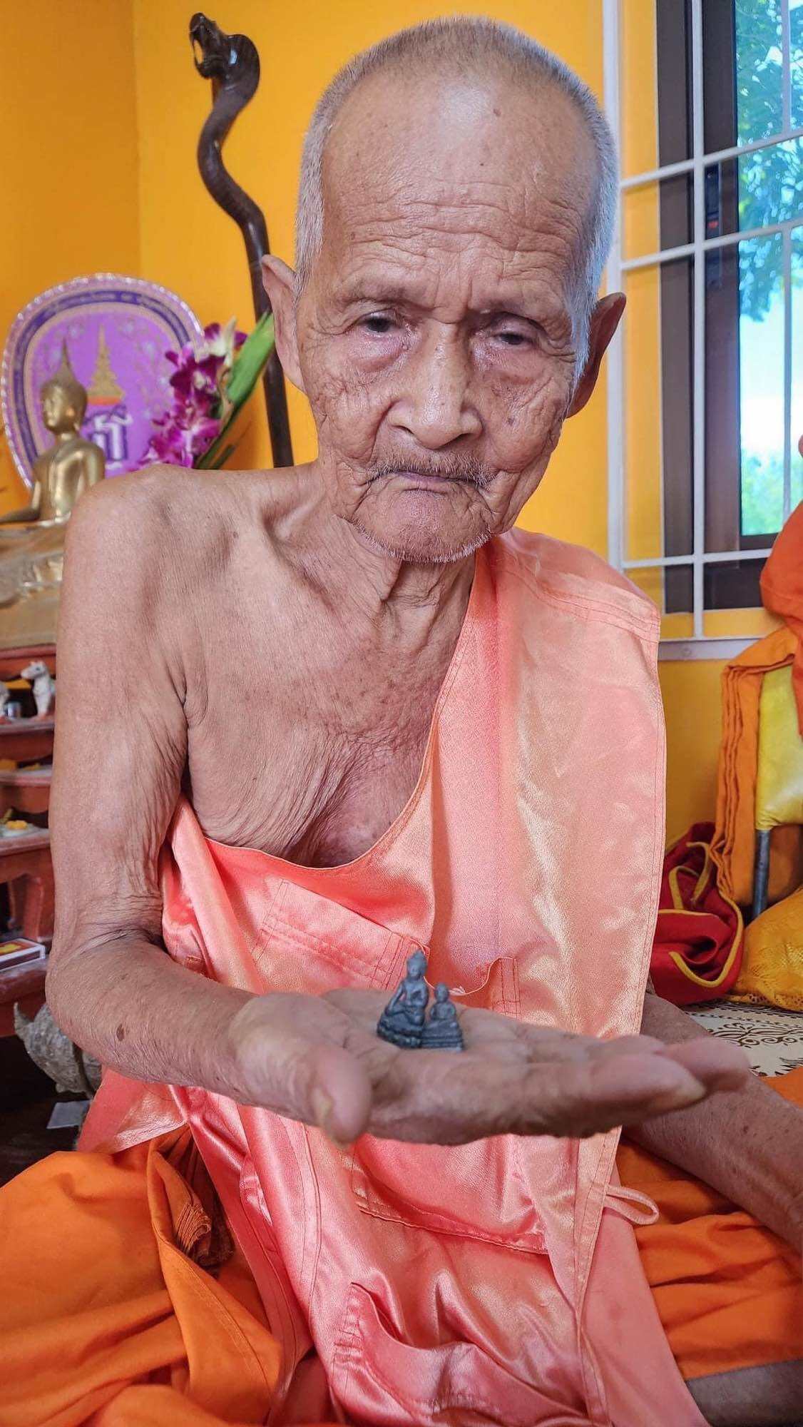 Elder Thai monk in orange robe holding Mongkol Hemarat Kring spiritual charm amulet in temple setting