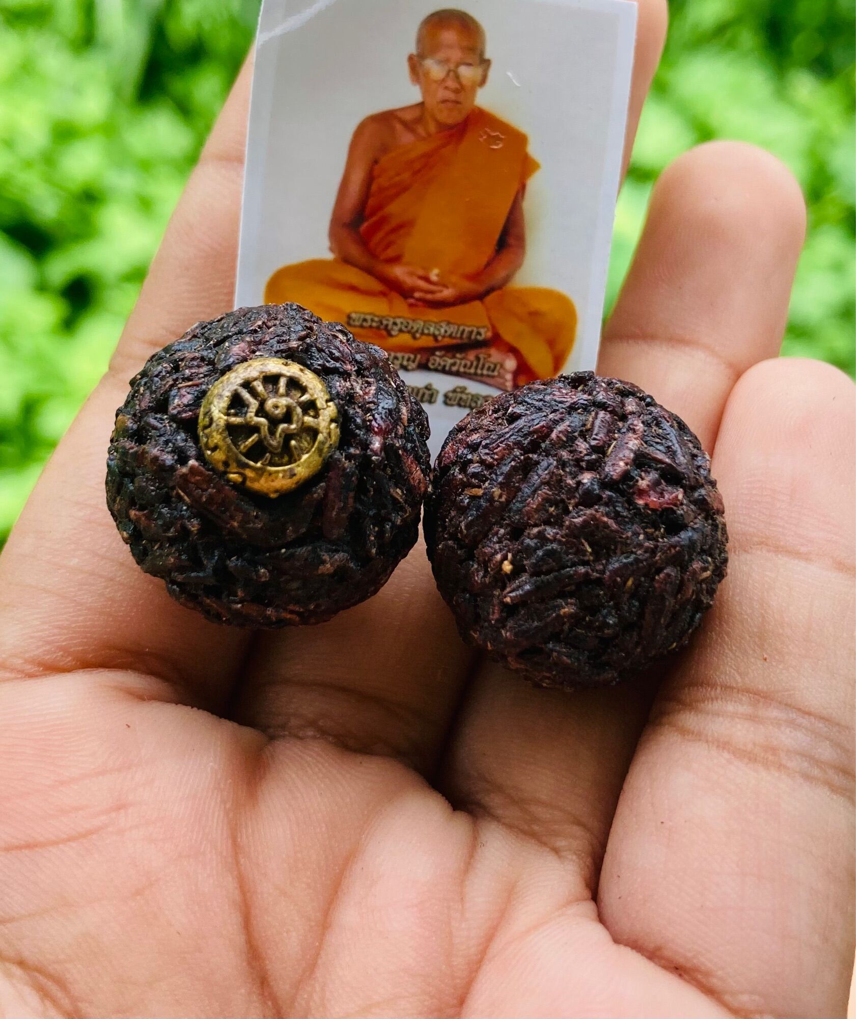 Hand holding two sacred black sticky rice bead amulet balls one with brass medallion and monk photo card