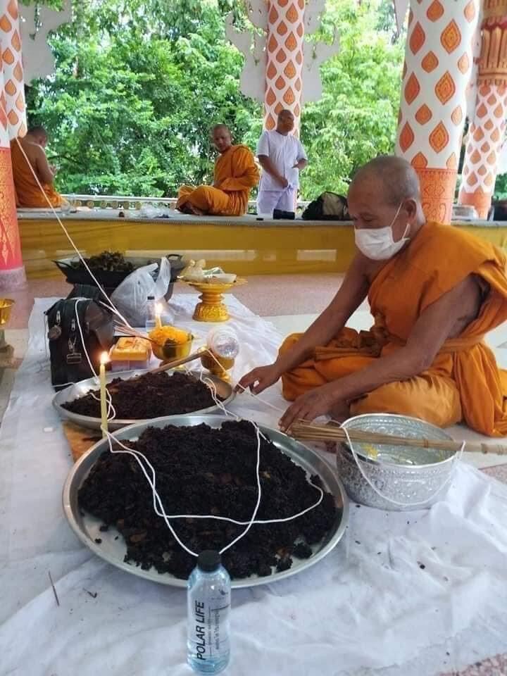 Masked Thai monk preparing sacred black sticky rice bead amulet on silver trays with temple offerings