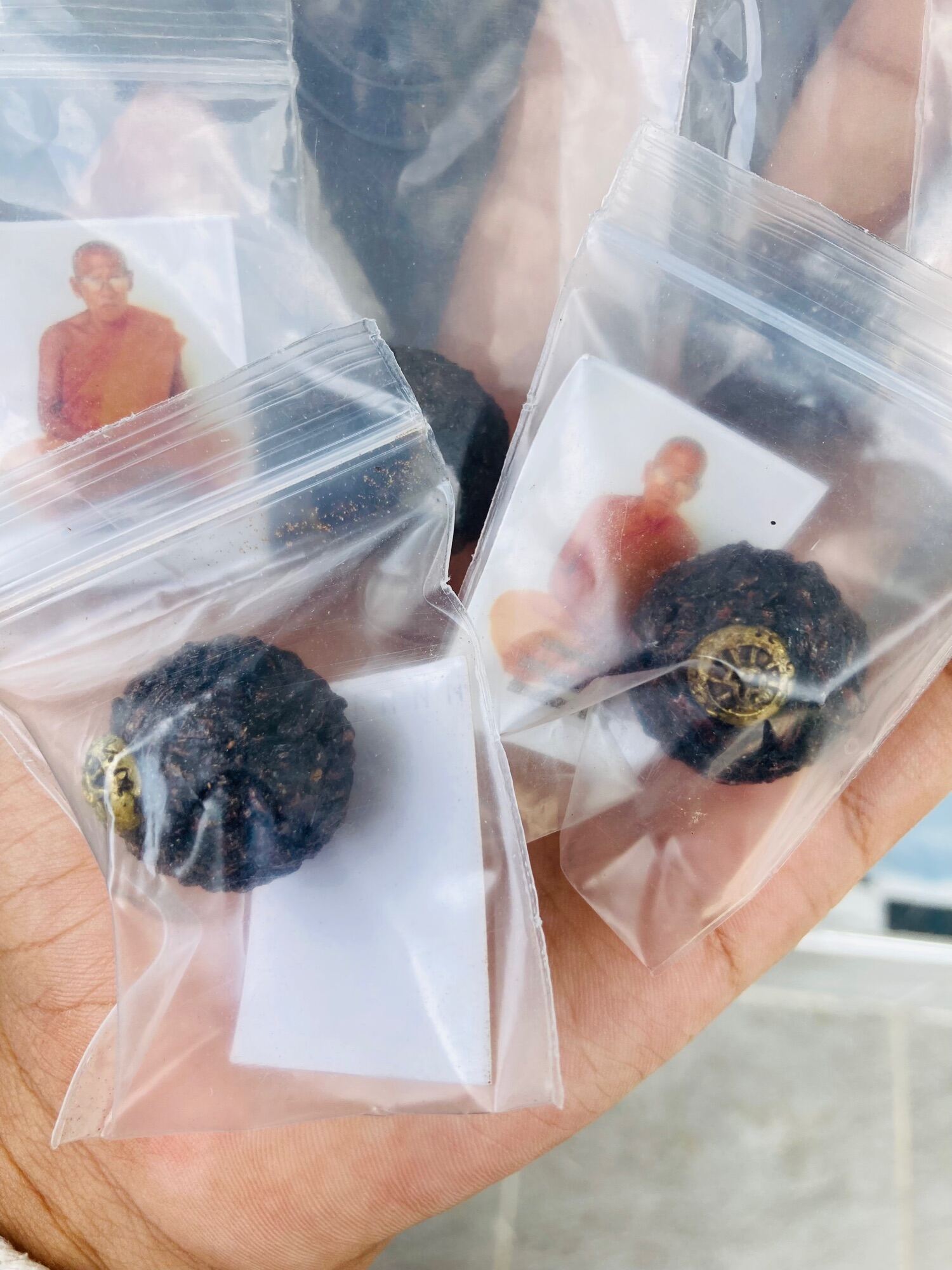 Hand holding small sealed bags with sacred black sticky rice bead amulet and monk photo cards