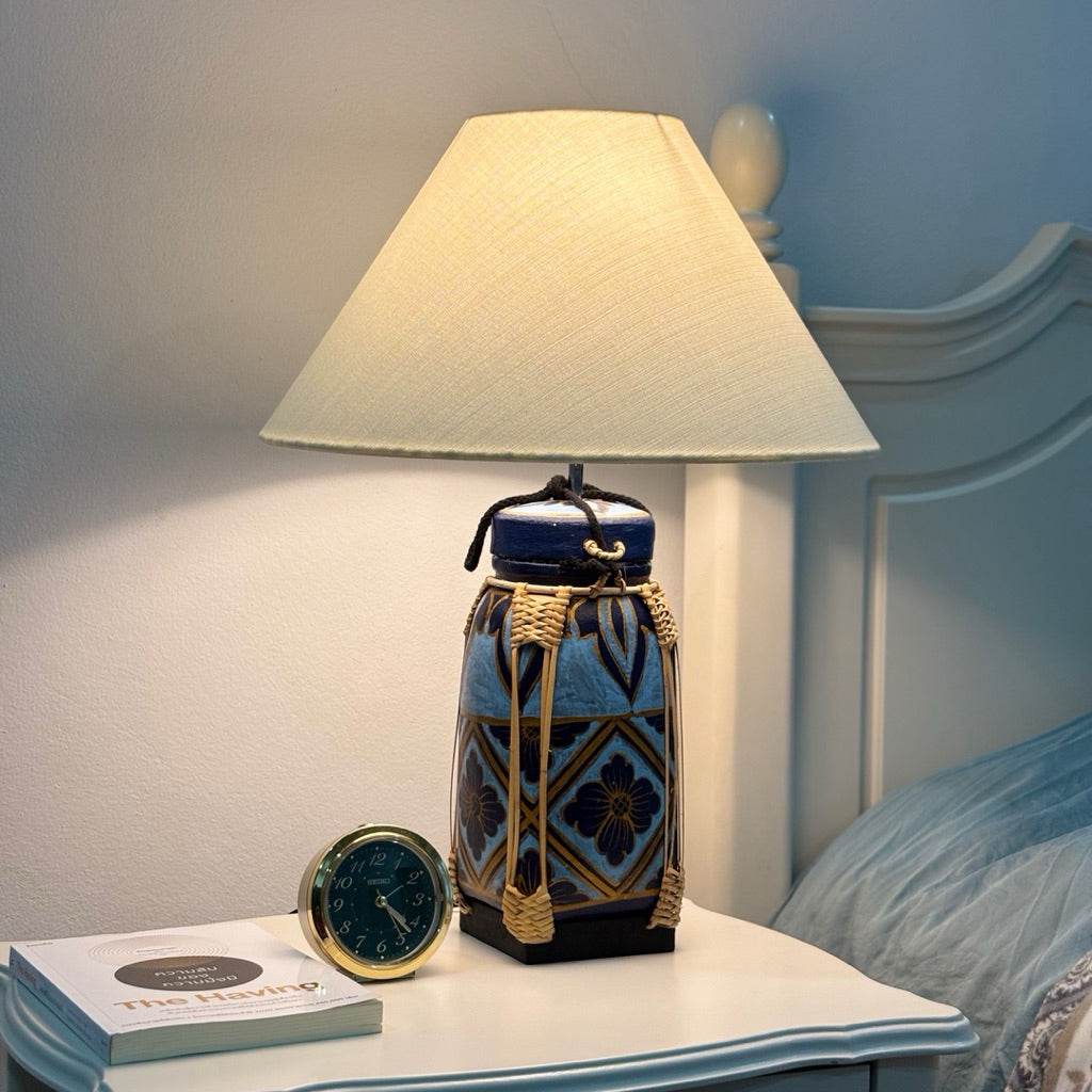 Woven bamboo rice basket lamp with a fabric shade placed on a bedside table next to a clock and book